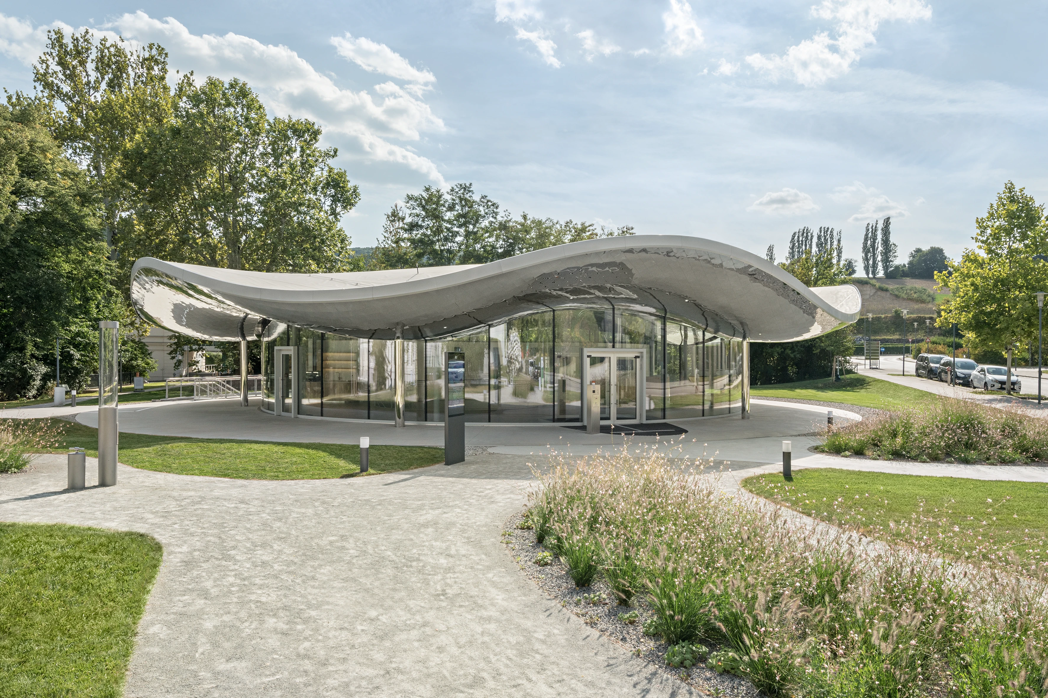 Das Bild zeigt ein Glashaus mit reflektierendem Dach in einer Parklandschaft. Foto von Hertha Hurnaus.